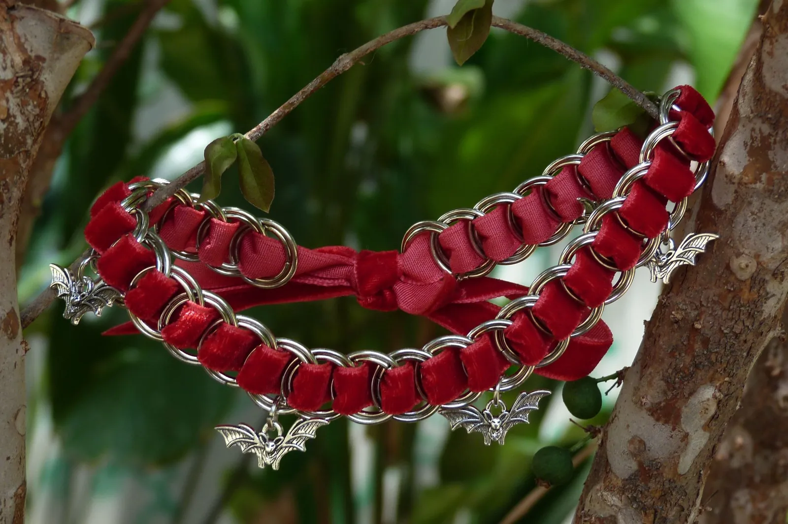 Gargantilla red bat de Liminal: terciopelo rojo entrelazado con aros metálicos y dijes de murciélago, fotografiada entre ramas.